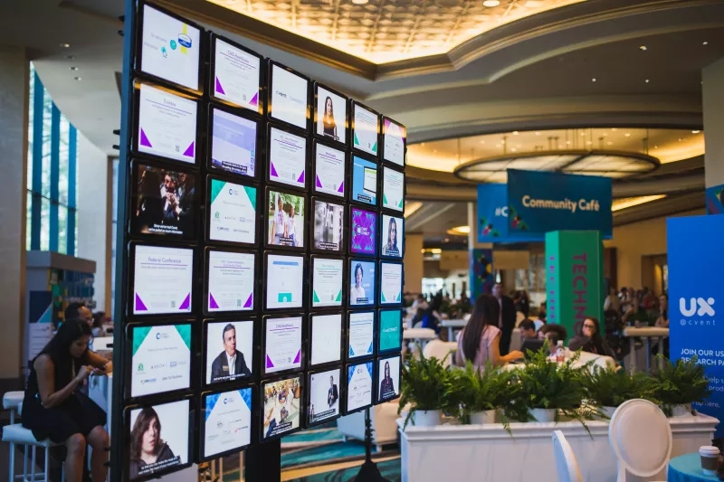 A wall of screens at a conference with people, plants, banners, and lights in the background.