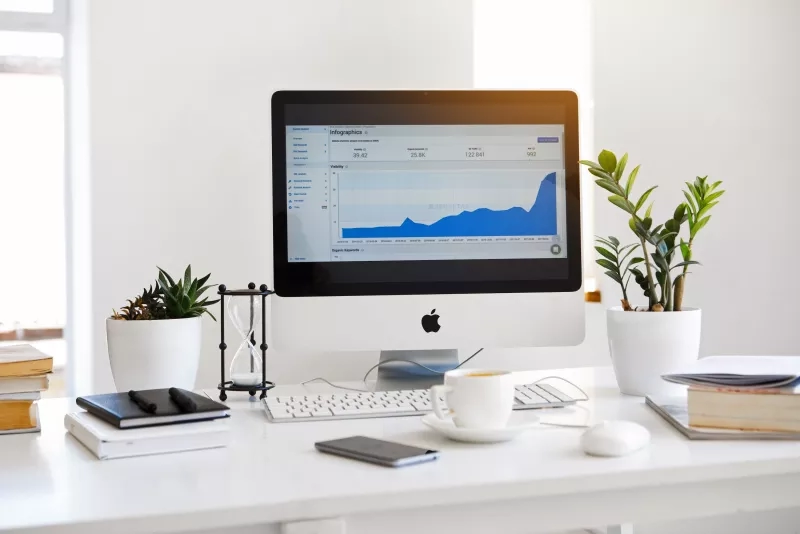 A white desk with an Apple monitor, keyboard, mouse, coffee, and potted plants on it.
