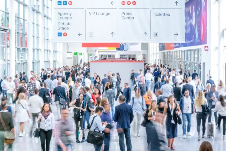 Blurry image of many people walking in a room with signs that read Agency Lounge, VIP Lounge, and Press Center.
