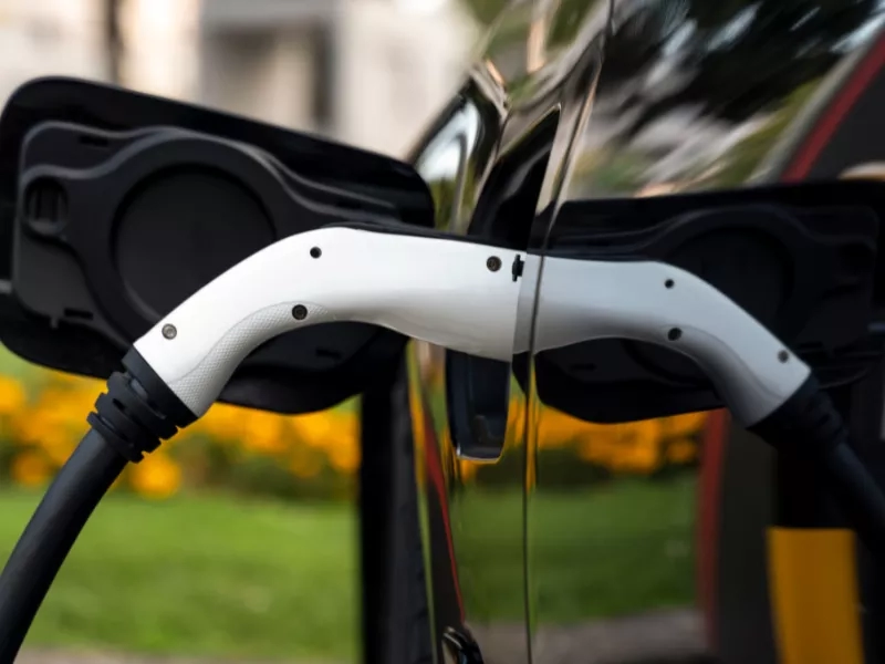 A white electric car charging connector plugged into a black charging port in an outdoor setting.